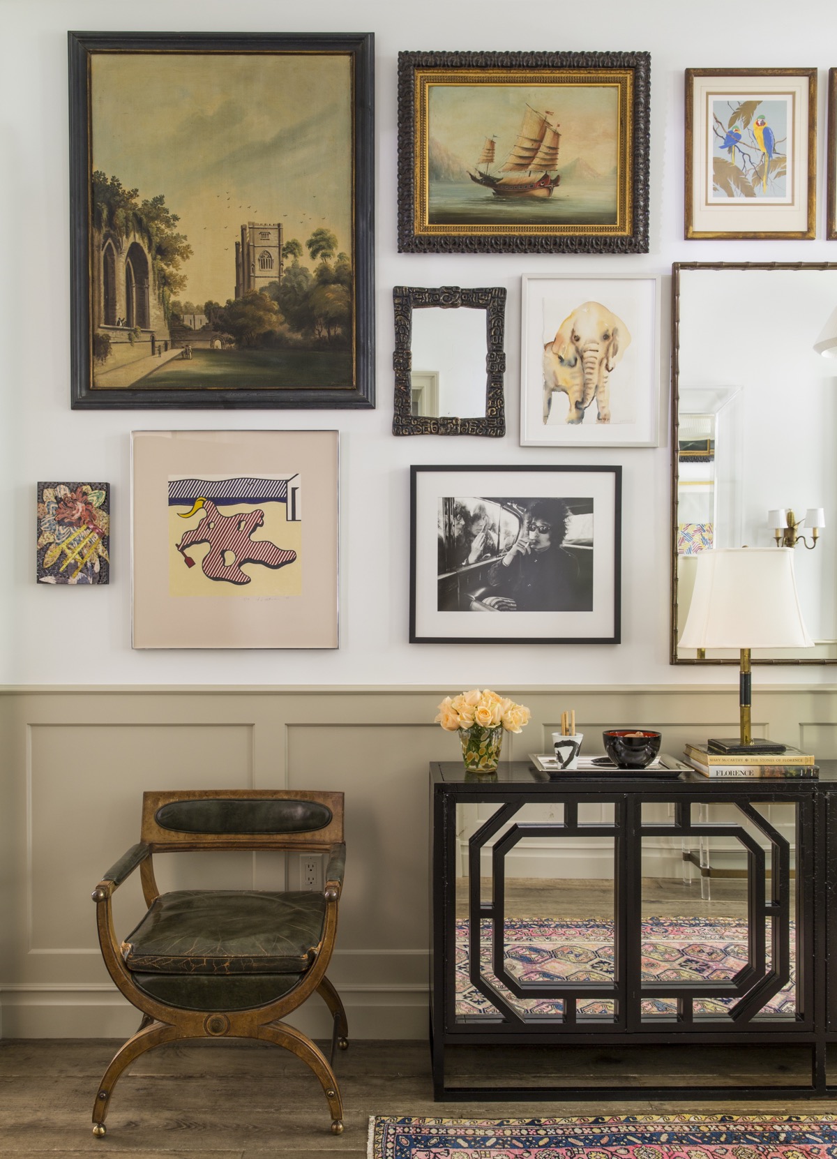 Entry hall with wood and leather chair, dark wood wall table with mirrored front, lamps, flowers and tray in front of light wall with various mirrors, paintings and photos.