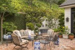 Exterior brick patio with iron frame chairs, sofa and table with glass top, patterned upholstered pillows, ivy climbing back wall, various plastered and potted trees and shrubs, grey metal fountain on far wall.