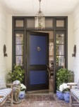 Front door overhang with blue door, matching benches with patterned cushions, potted plants.brick walkway.