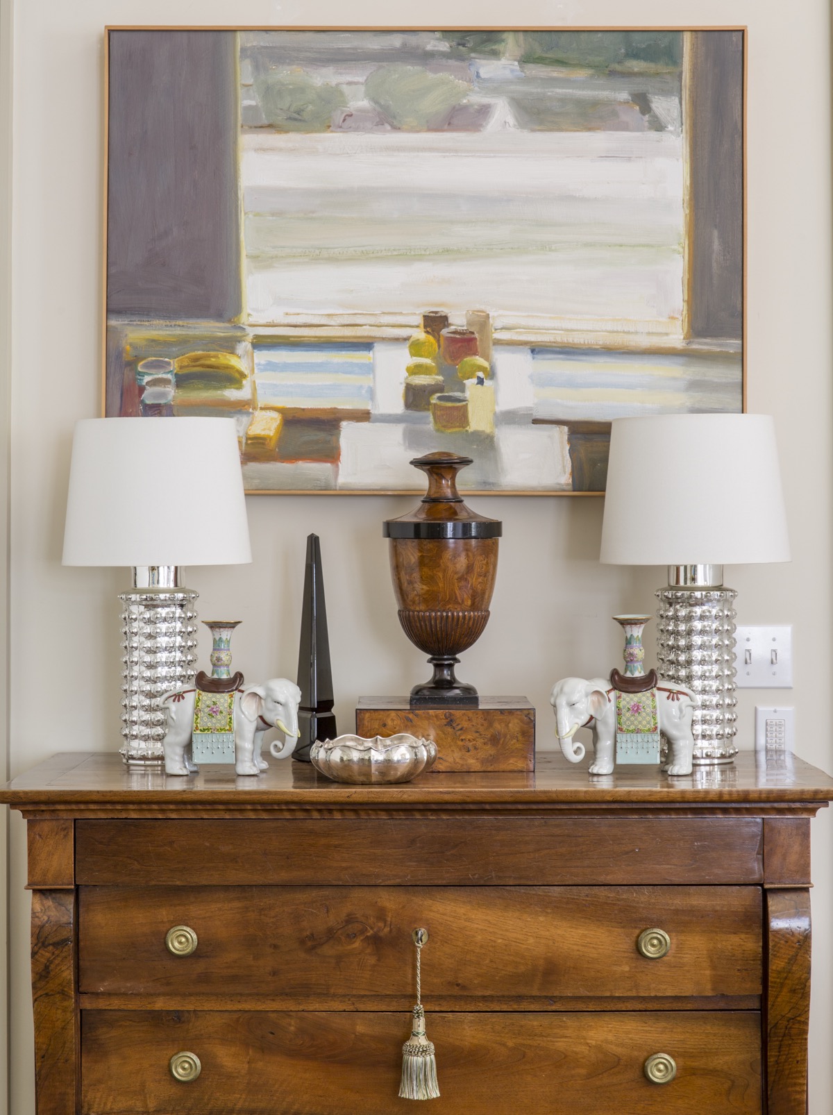 Detail of wood dresser top with pair of silver lamps, ceramic elephants, bowl and urn, in front of light wall with painting.